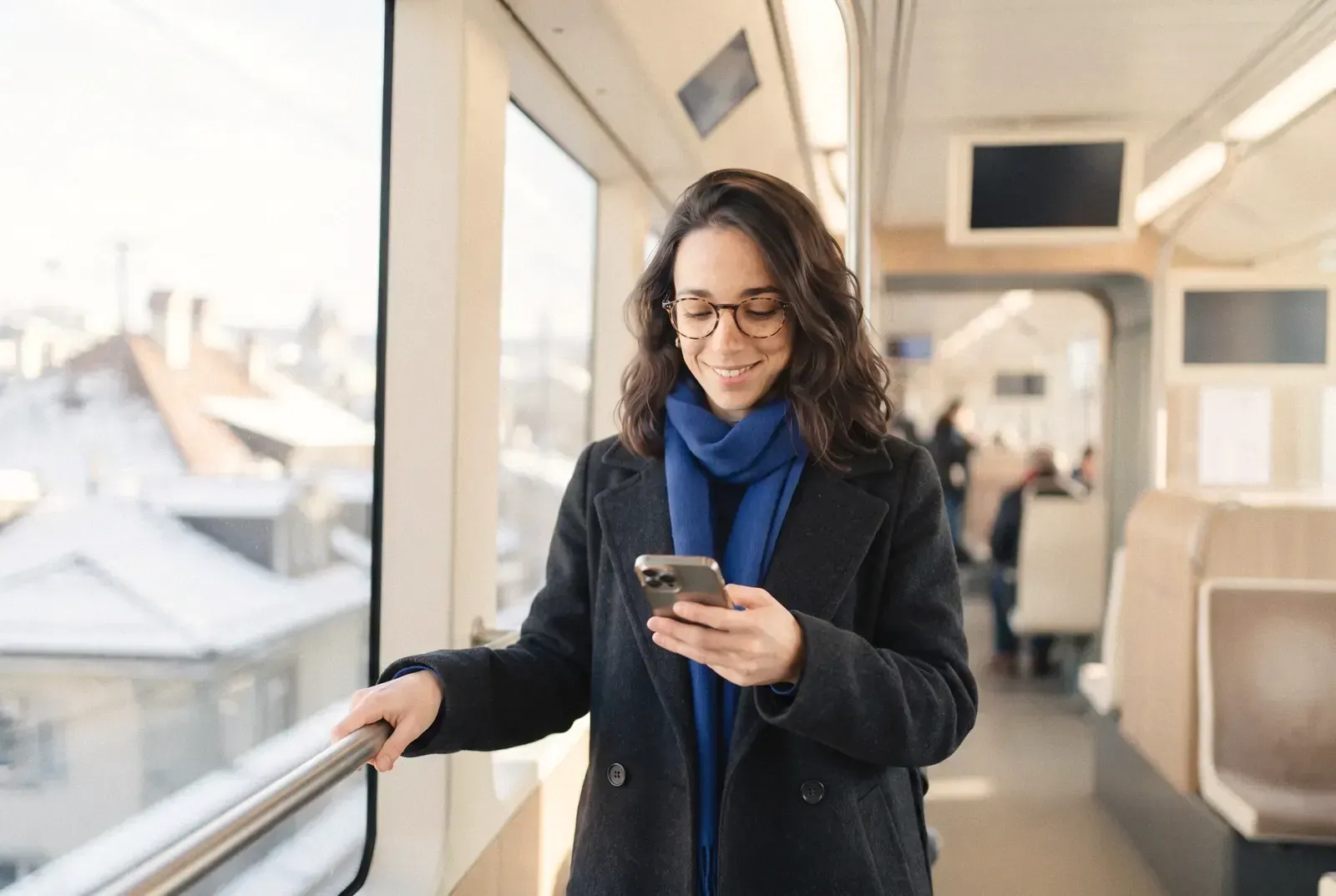 Une jeune femme souriante consulte son téléphone debout dans un tram, la ville enneigée derrière la vitre