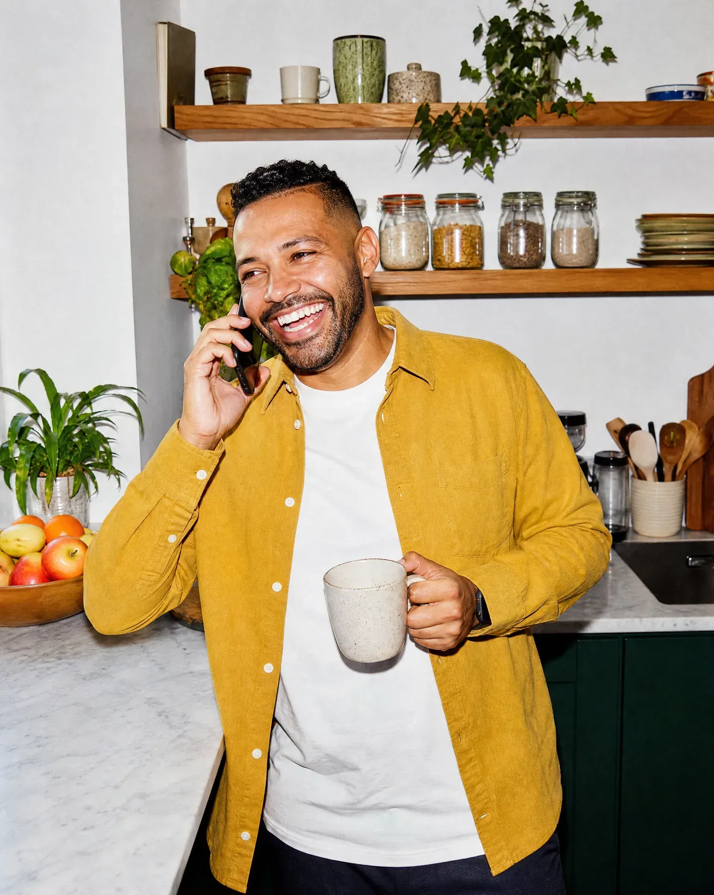 Un homme en surchemise moutarde rit au téléphone dans sa cuisine, une tasse à la main.