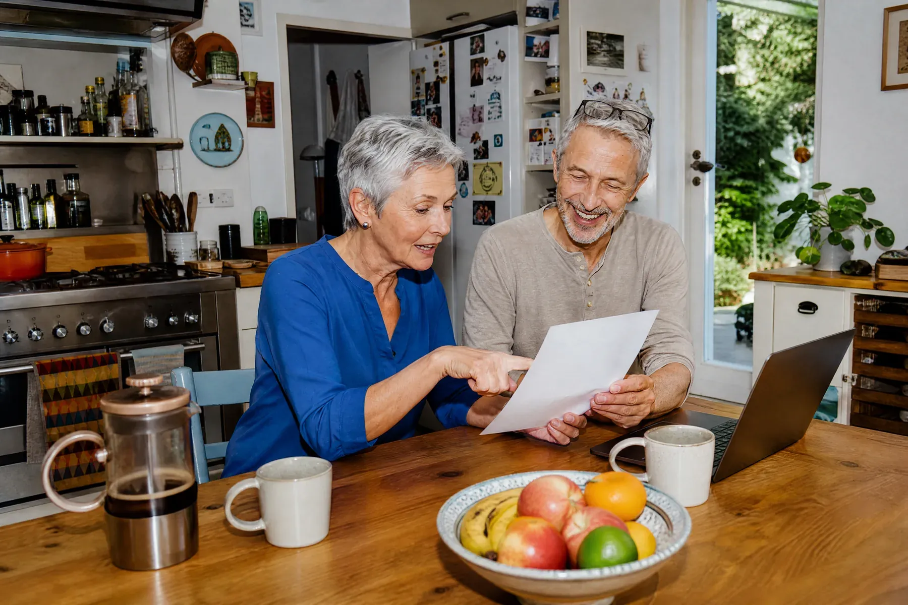 Un couple aux cheveux gris compare une feuille à la table de la cuisine, à côté d’un ordinateur portable et d’une coupe de fruits